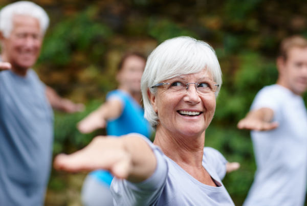 Smiling elderly woman with gray hair and glasses doing outdoor group exercise, extending arms forward.