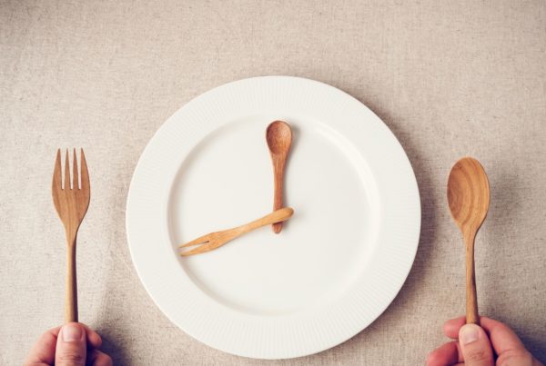 Two wooden utensils lie on a white plate like the hands of a clock to represent intermittent fasting. A fork and spoon sit on either side.