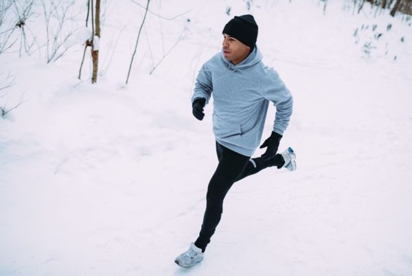 From above, a man runs in the snow wearing a black hat and grey shirt.