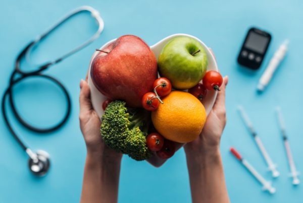 Two hands hold fruit in a heart-shaped bowl over a blue background holding medical equipment for treating metabolic disease.