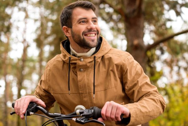 A bearded man smiles and rides his bike on a wooded path, representing the positive momentum he’s gaining in his health.