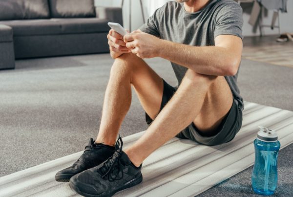 A man sits on a yoga mat in his living room, holding his phone after finding time to workout.