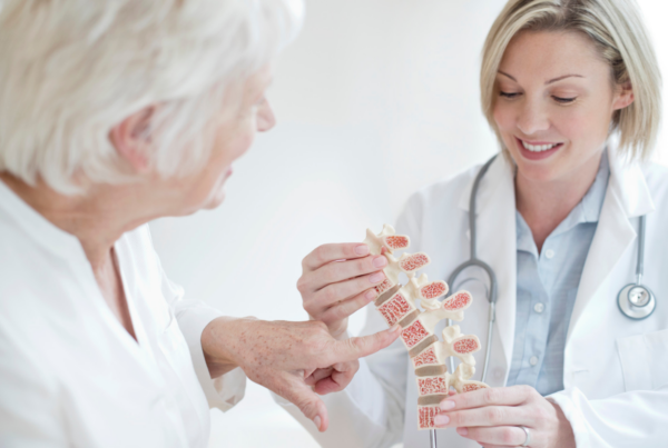 An older woman discusses her risk factors for osteoporosis with her physician, using an anatomical model of a split spine.