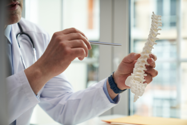A doctor points to a small model of a spine and pelvis after his patient asks him, “Can osteoporosis kill you?”