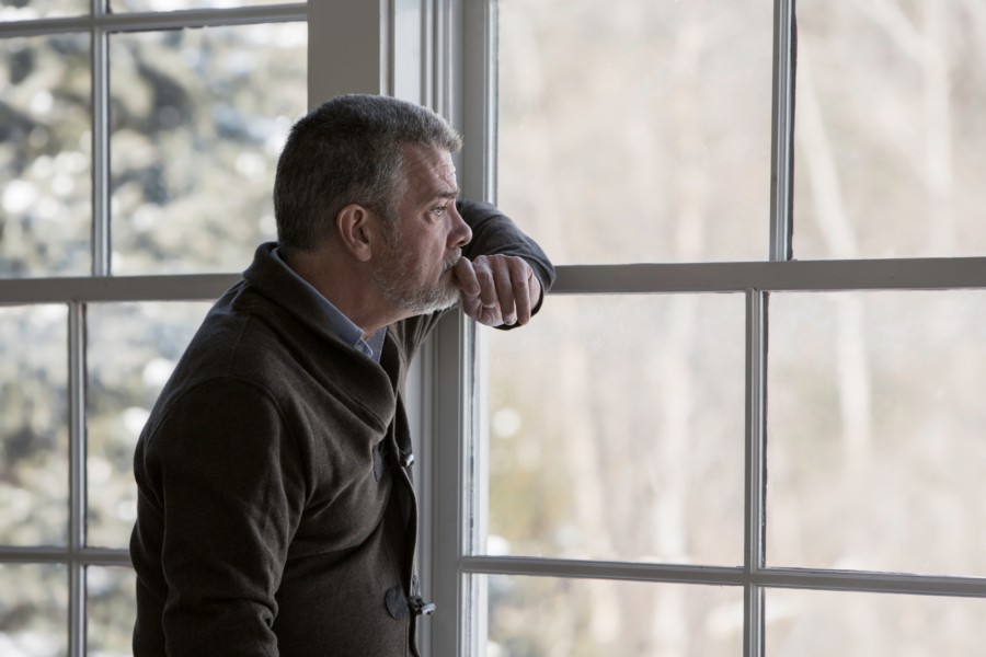 A man leans and stares out of a window, grieving after loss.