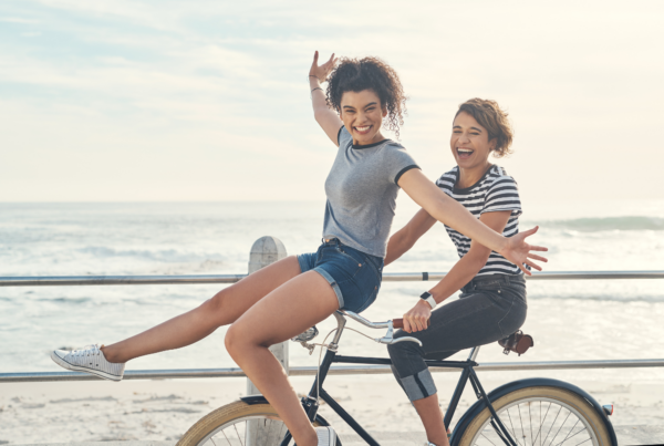 A pair of friends ride a bike together near the ocean, demonstrating living with a zest for life.