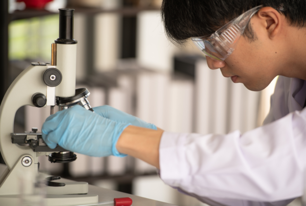 A scientist in protective goggles and gloves closely examines a sample under a microscope in a laboratory setting.