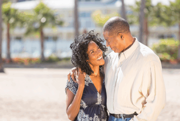 Smiling couple embracing on a sunny beach with palm trees and a resort building in the background.