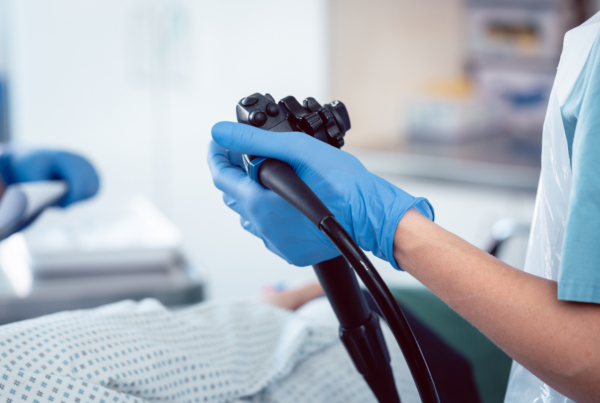 A clinician holds an endoscope before a colonoscopy procedure, with the patient prepped on the exam table.
