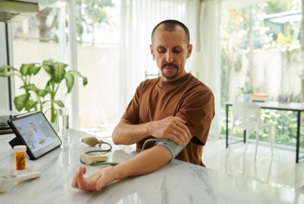 Man checks his blood pressure with a home arm cuff at his kitchen counter during a video call with his doctor.
